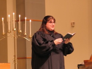 Katie, in a black alb, preaching from a small worship book